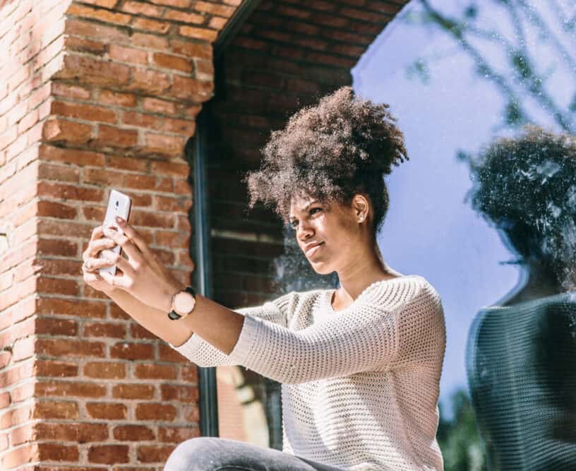 How Do Hair Extensions Work on Short, Straight, Curly, or Thinning Hair 10 Beautiful young African American girl with a joyful expression, taking a selfie next to a large window and looking at herself in the reflection.