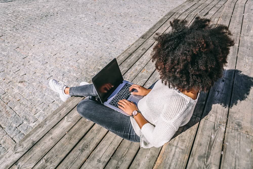 Brunette woman with an afro wearing hair extensions, casual clothes, and sunglasses while working on a laptop while enjoying the sunlight during a sunny day.