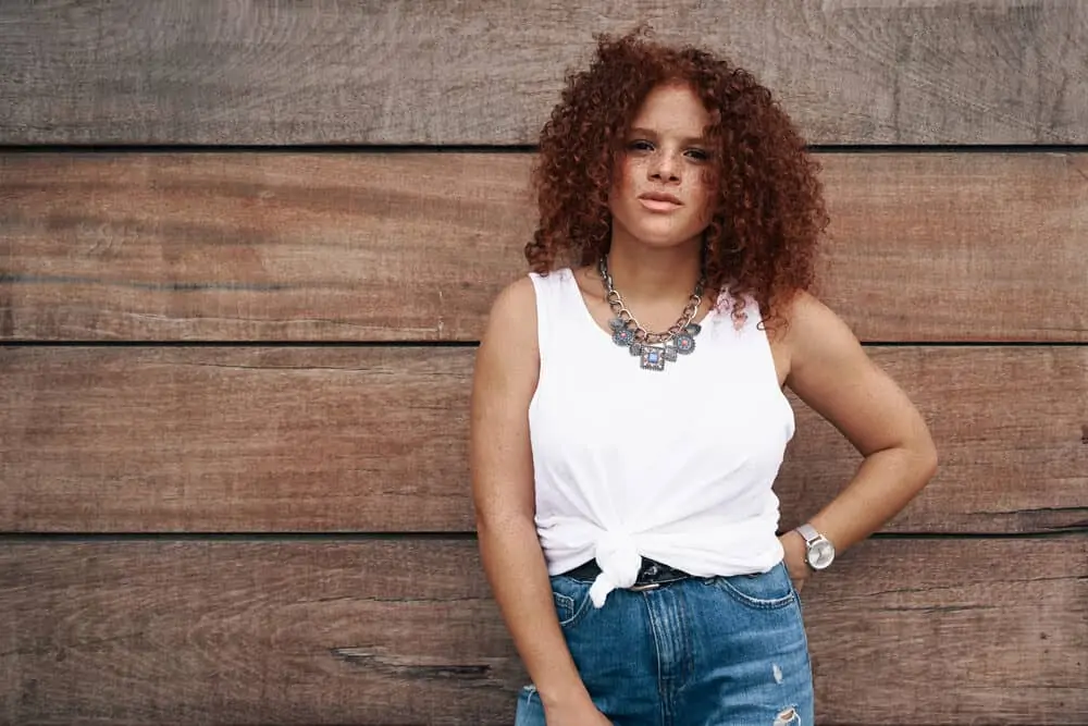 How to Neutralize Red Tones in Hair: Fix Your Hair at Home 1 A woman in casual clothes is standing on the wooden background. She has curly red hair with orange undertones and freckles across her face.