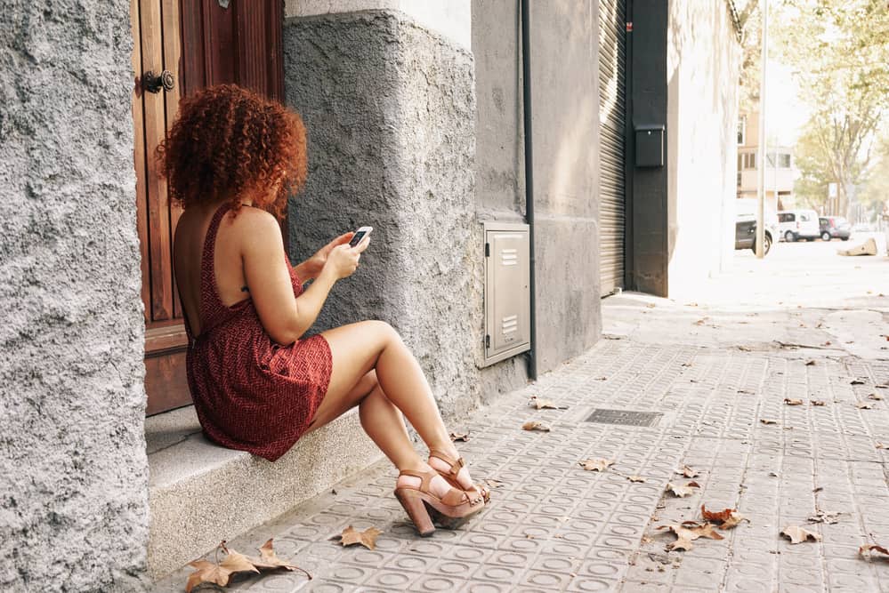 A woman with red pigments in her hair wearing a dress and sandals sits on the sidewalk beneath a tree, talking on her smartphone.