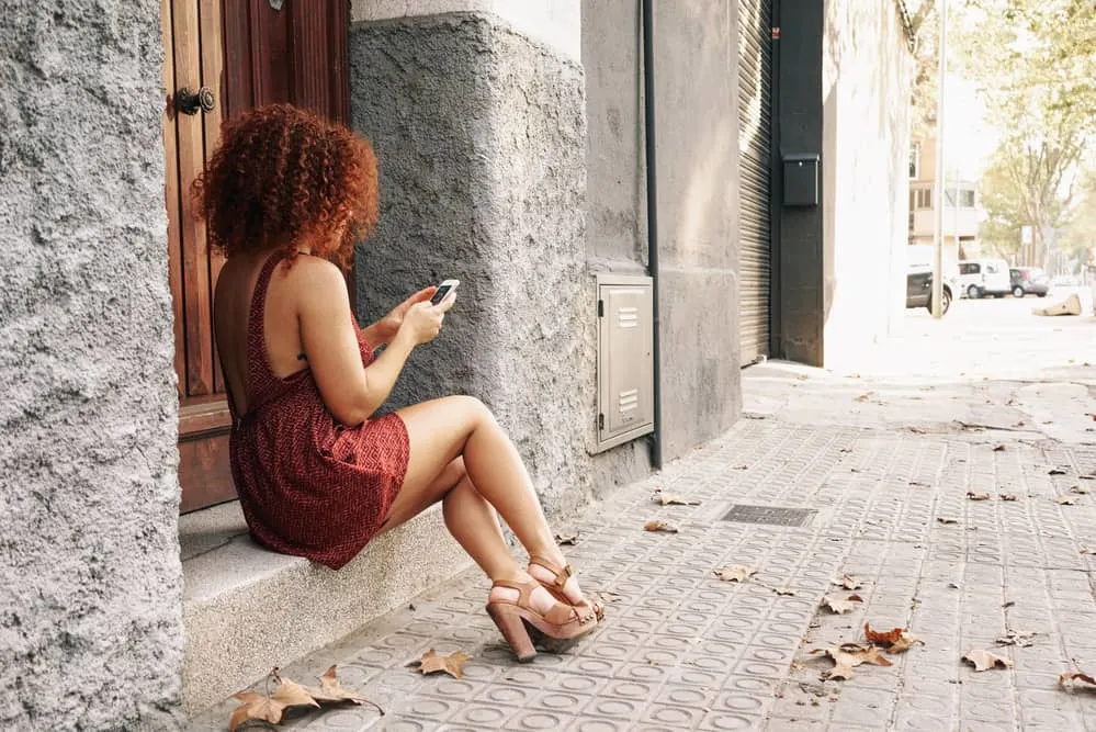 A woman with red pigments in her hair wearing a dress and sandals sits on the sidewalk beneath a tree, talking on her smartphone.