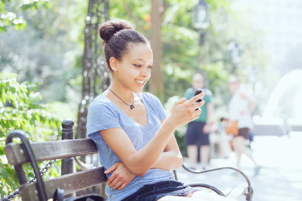 A young black woman with head lice is sitting on a park bench while using an iPhone 12 with naturally curly hair.