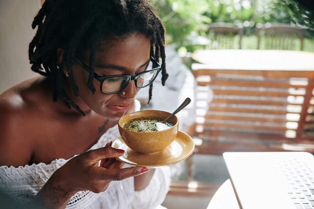 African female wearing dreads and eyeglasses while eating a bowl of soup at an outdoor eatery.