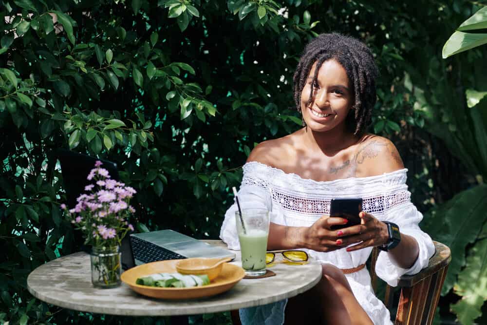 Happy female sitting at the table with her laptop and mobile phone with green plants in the background.