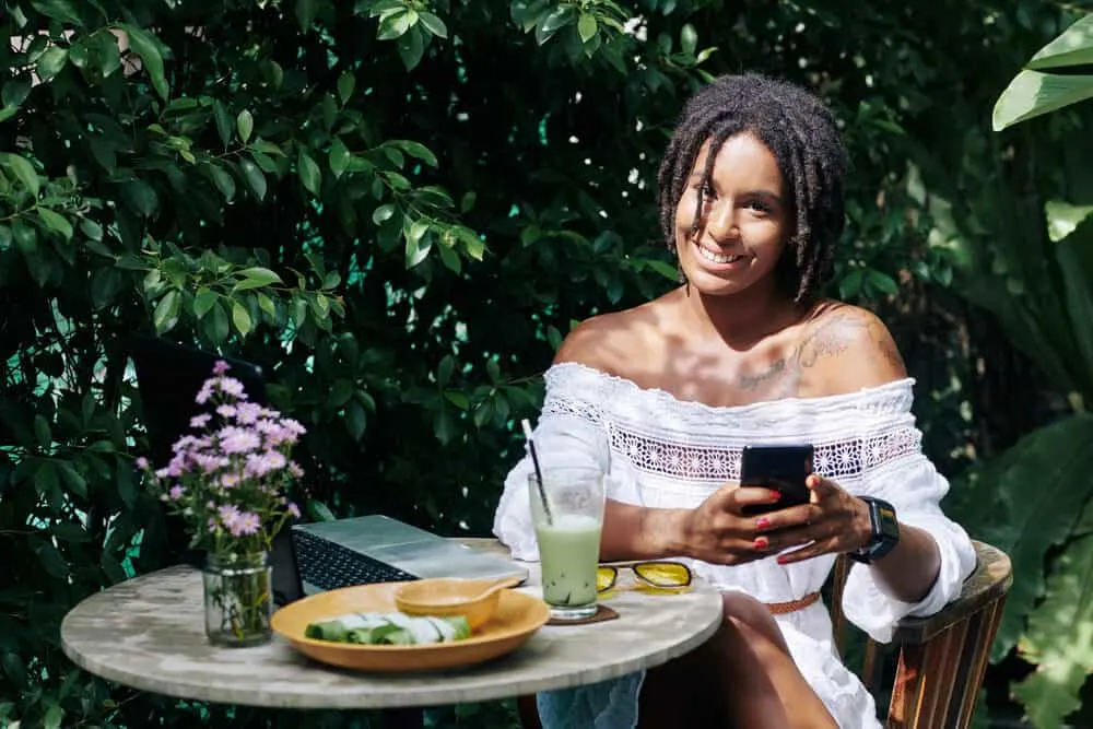 Do Dreads Smell? How to Stop Dreadlocks From Smelling 4 Happy female sitting at the table with her laptop and mobile phone with green plants in the background.