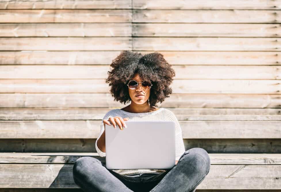 A young brunette girl with curly ombre thicker hair stares at her laptop as the sun shines on her face.