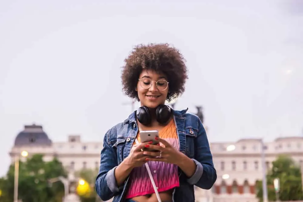 A woman with dark skin wearing black headphones smiles at her smartphone on the street wearing a shirt with vibrant colors.