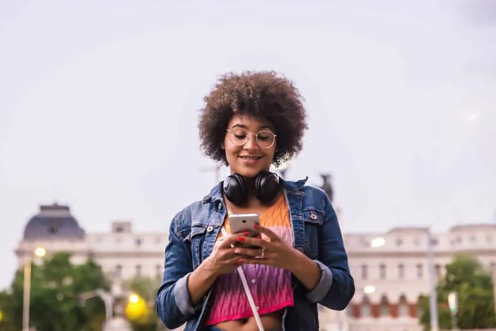 What Color Can I Dye My Black Hair Without Bleaching? Hair Dye Options for Dark Hair Colors 5 A woman with dark skin wearing black headphones smiles at her smartphone on the street wearing a shirt with vibrant colors.