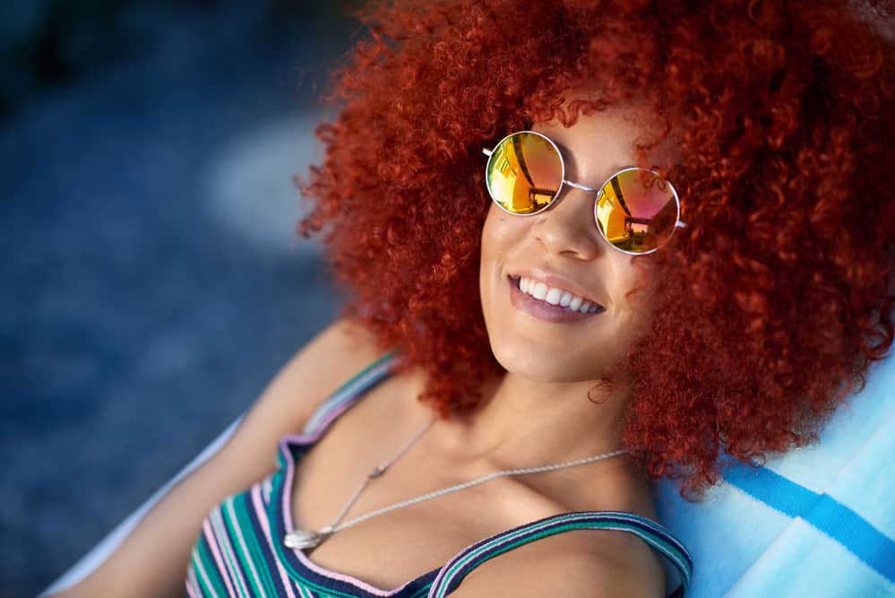 Black girl smiling wearing a silver necklace, shades, and curly red hair. 
