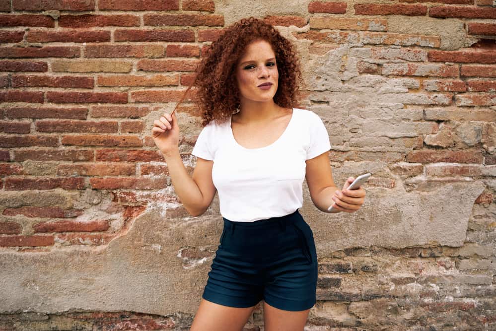 A woman, wearing a white t-shirt and black shorts with red brassy hair standing on the brick wall background.