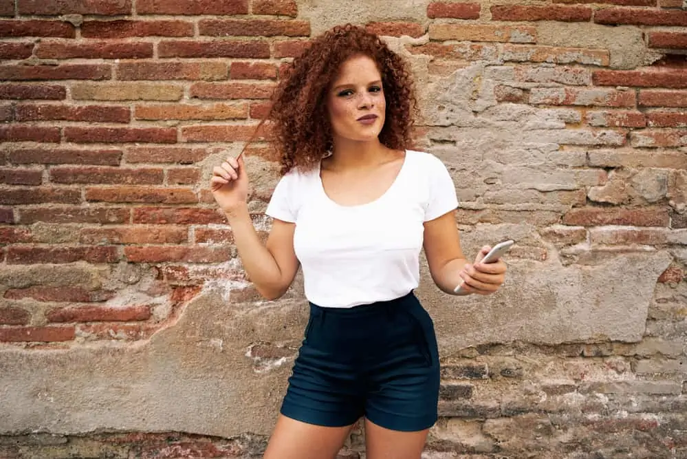 How to Neutralize Red Tones in Hair: Fix Your Hair at Home 2 A woman, wearing a white t-shirt and black shorts with red brassy hair standing on the brick wall background.