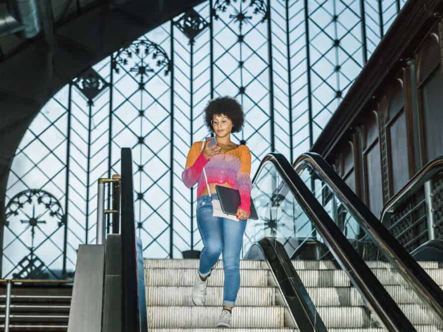 A woman with dark hair looks at her phone while going down the stairs inside a train station.