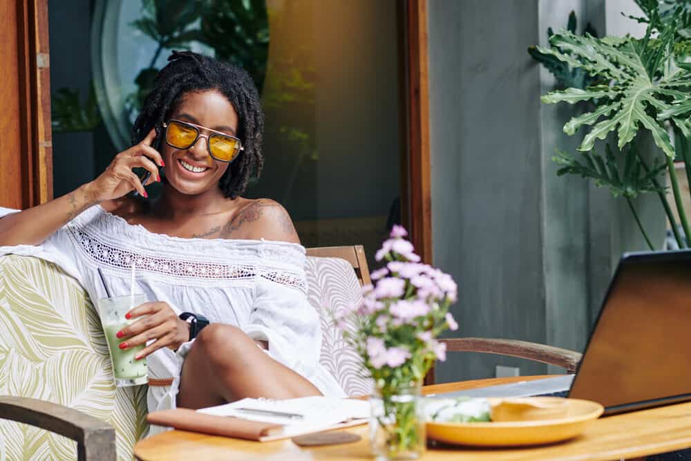 African American female sitting on a sofa outside drinking a green smoothie while talking on the phone.