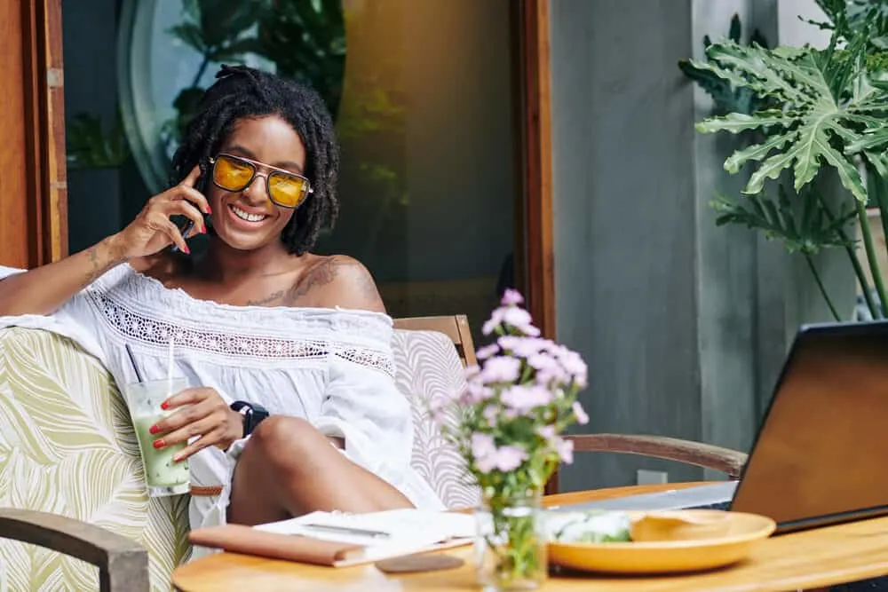 Do Dreads Smell? How to Stop Dreadlocks From Smelling 3 African American female sitting on a sofa outside drinking a green smoothie while talking on the phone.