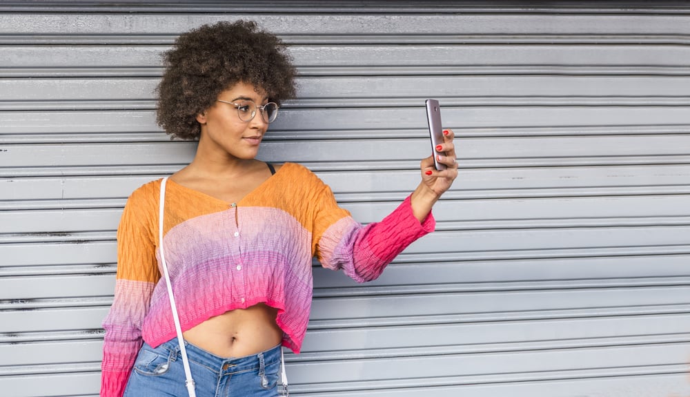 A pretty woman with glasses and a light-colored hair tone is dressed in colorful spring clothing next to a metal door.