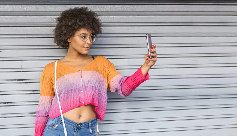 What Color Can I Dye My Black Hair Without Bleaching? Hair Dye Options for Dark Hair Colors 2 A pretty woman with glasses and a light-colored hair tone is dressed in colorful spring clothing next to a metal door.