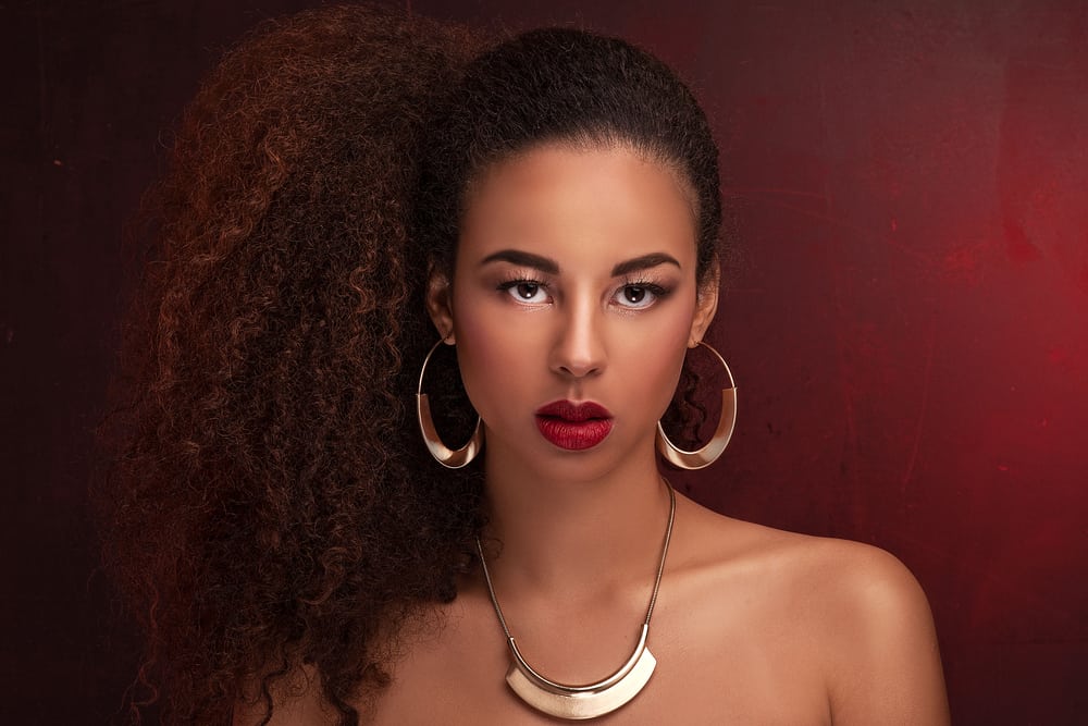  Elegante schwarze Frau mit goldenen Ohrringen und rotem Lippenstift, die Mikroring-Haarverlängerungen trägt, die an ihrem echten Haar befestigt sind.