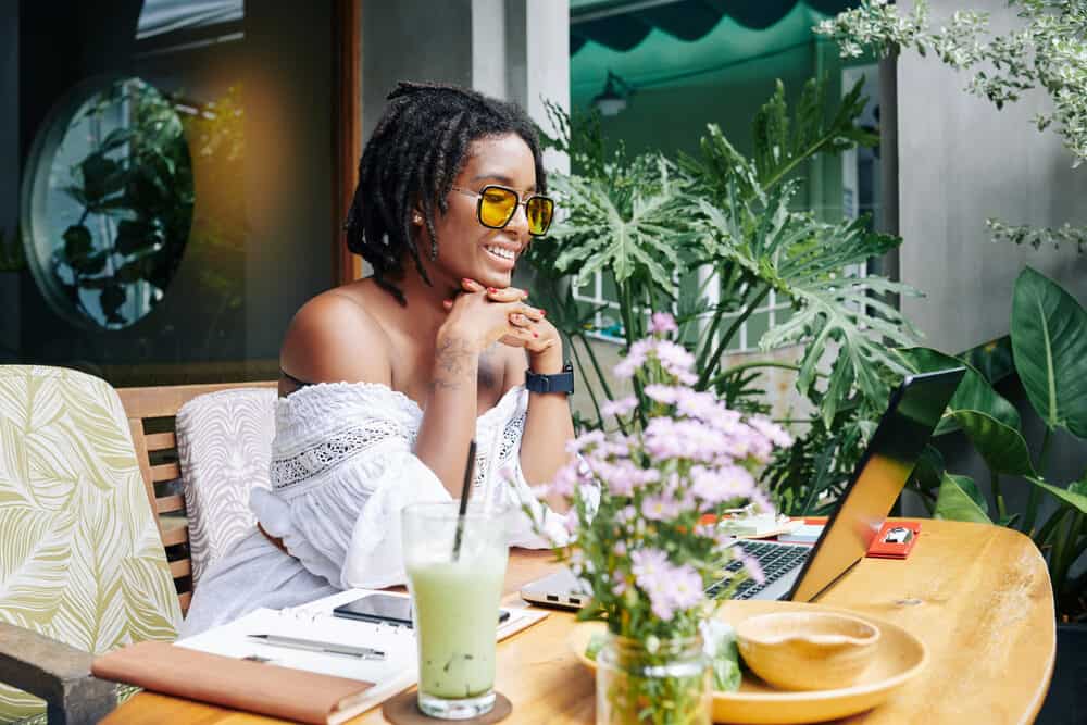 Lady with curly locs sitting at a table wearing glasses while using a laptop.