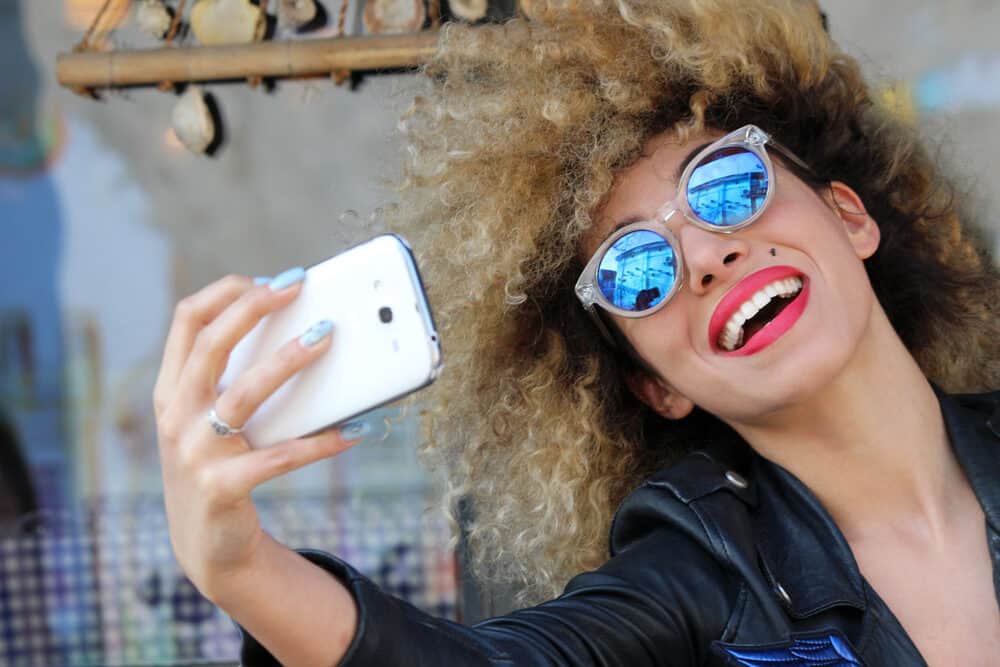 Mixed female smiling while taking a selfie wearing curly hair strands with brassy undertones.