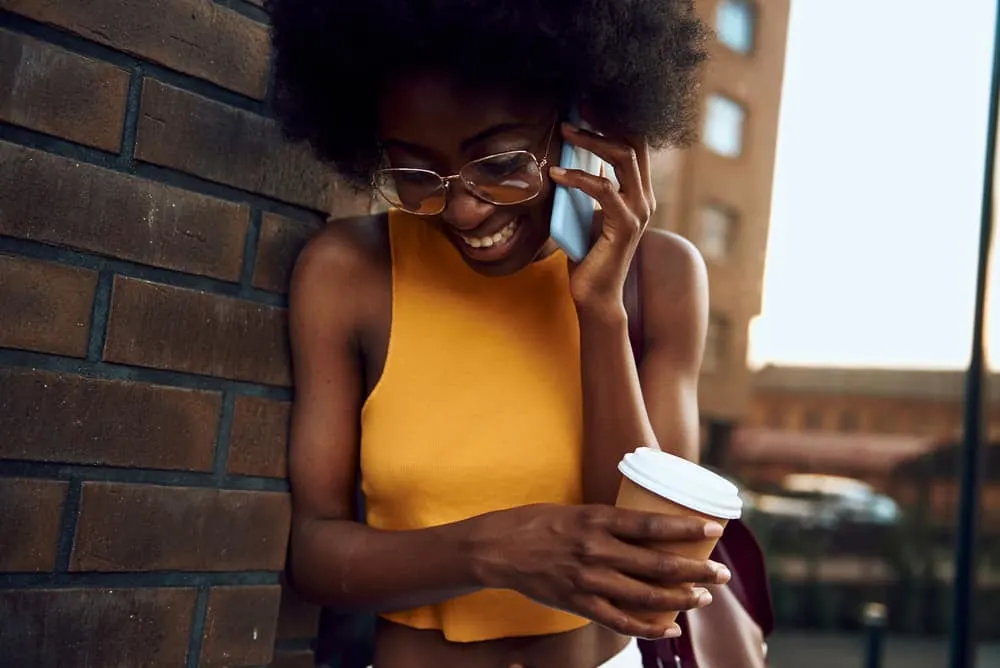 Best Blow Dryer for 4C Hair: Top Dryers for Women with Kinky Curly Natural Hair 7 A smiling lady standing near a brick wall with a coffee while talking on her phone.