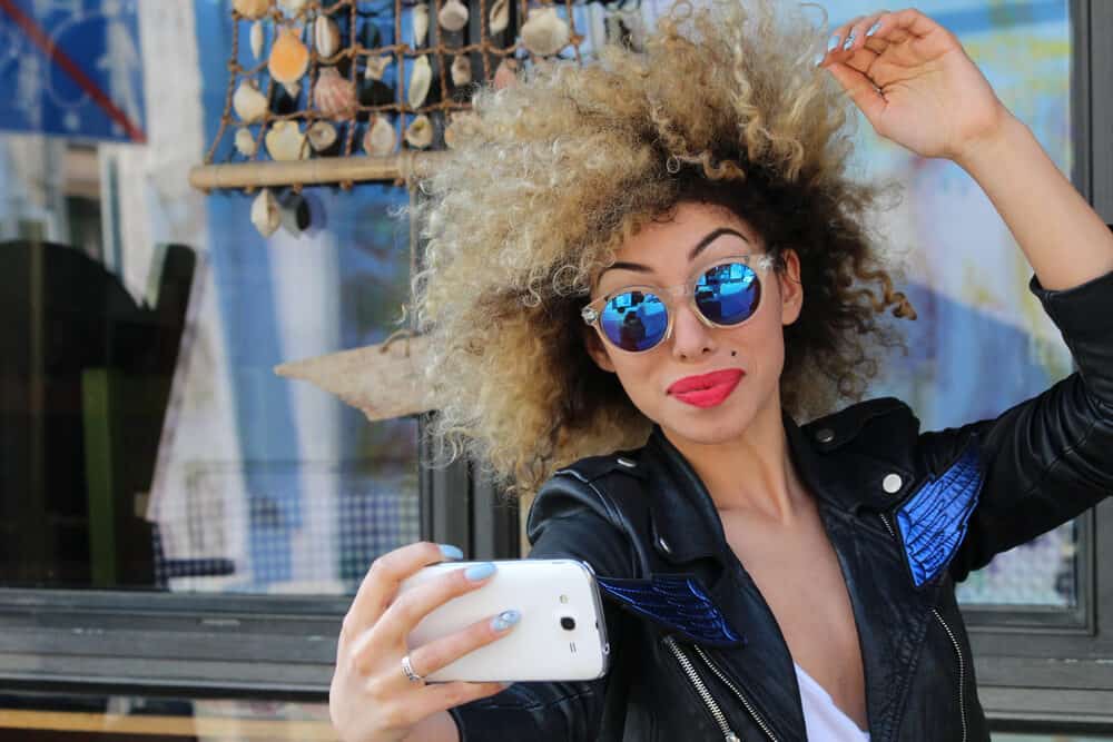 Black girl wearing a leather jacket, delicate shades and red lipstick, with a pale yellow shades over ash brown hair.