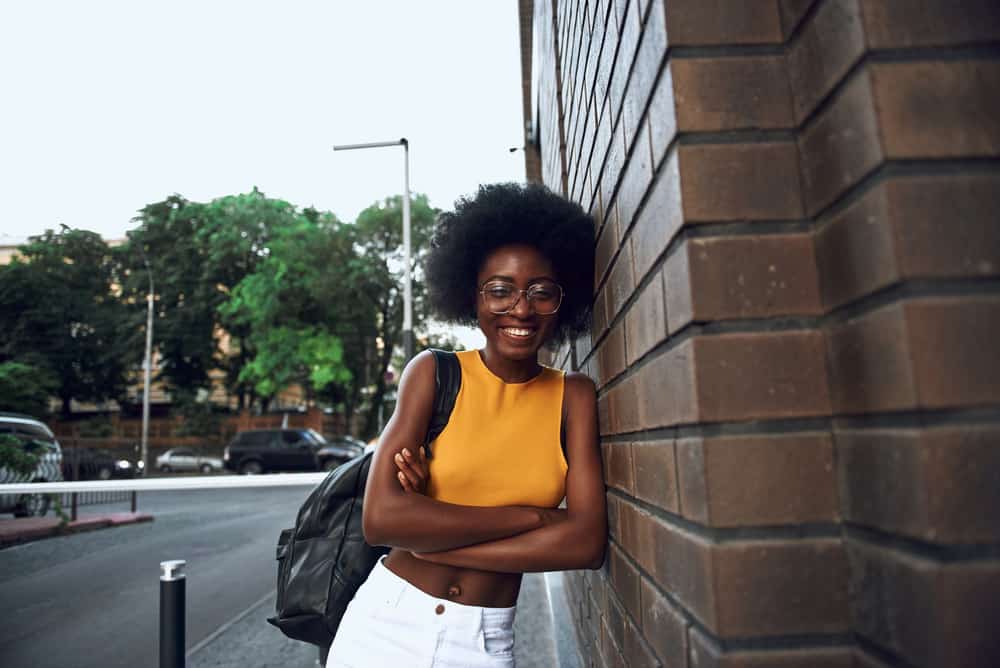 African American woman with 4C natural hair and a cheerful demeanor wearing a black handbag and standing near a brick wall with her arms crossed.