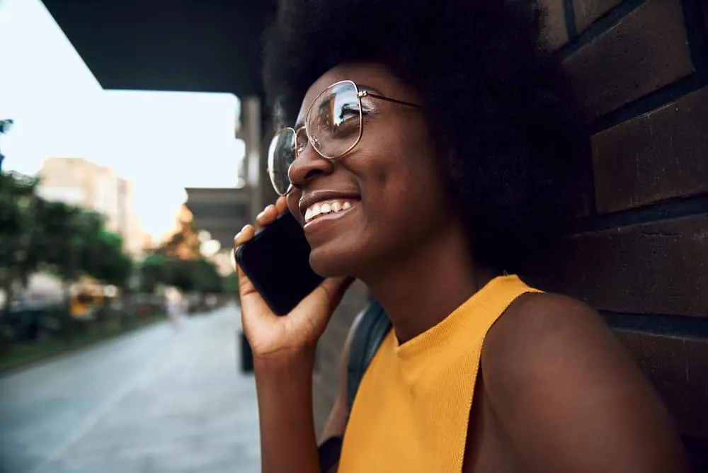 Best Blow Dryer for 4C Hair: Top Dryers for Women with Kinky Curly Natural Hair 5 Cute black woman with naturally curly hair in glasses talking on the phone while leaning against a brick wall.