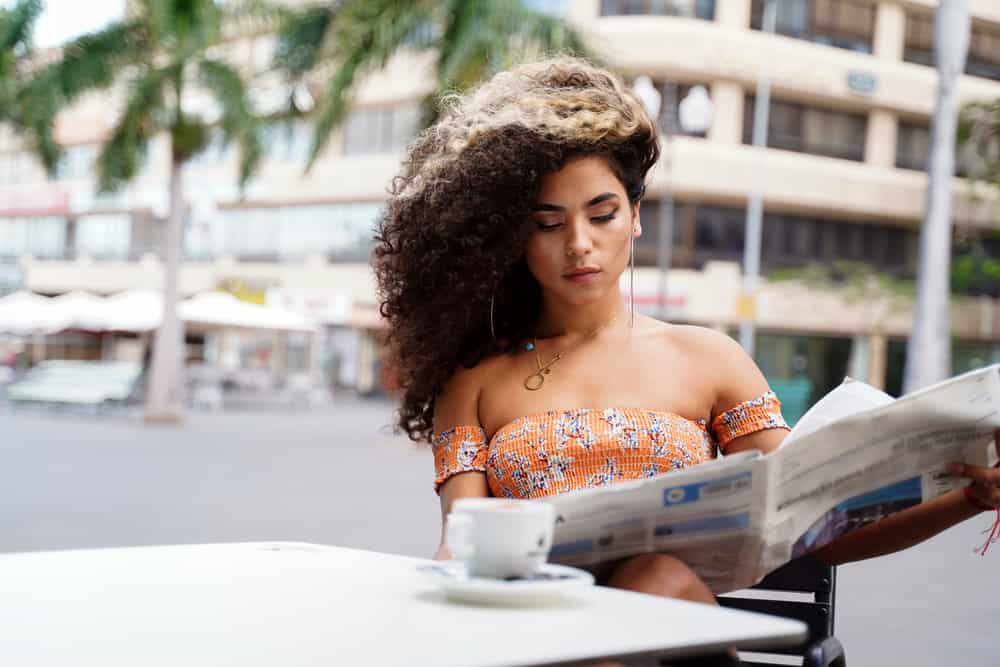  Mujer hermosa que lee un periódico, mientras usa aretes de aro y bebe una bebida líquida suave.