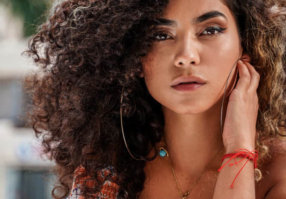  Femme colombienne aux cheveux ondulés de type 3 et à la peau caramel portant un maquillage neutre et un spray capillaire brillant.