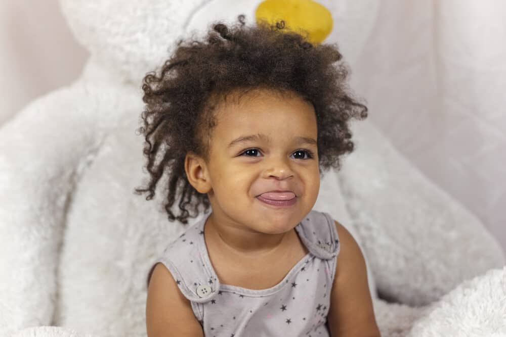 A lovely little baby girl with curly hair sitting on a huge, plush teddy bear playing with homemade slime