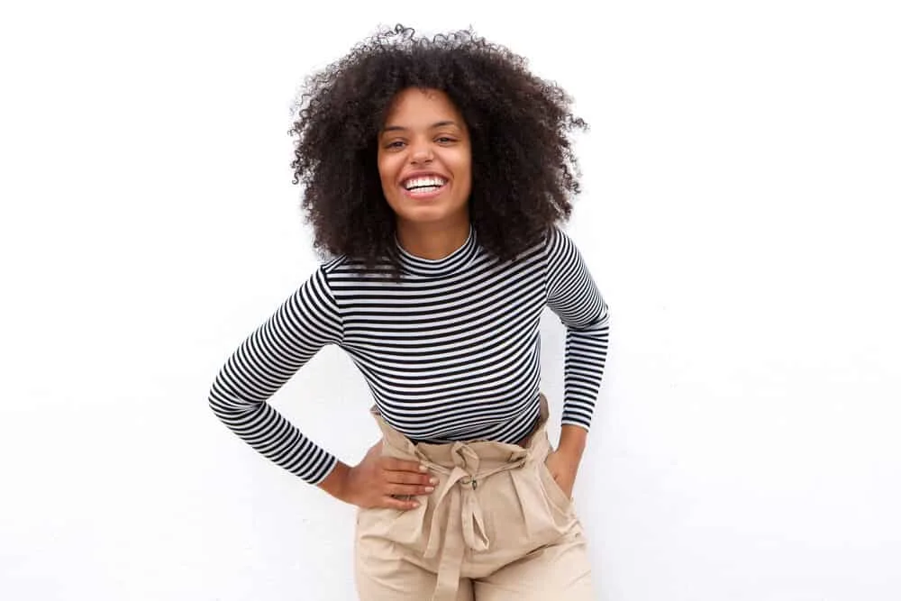 African American female with an even length hairstyle in its curly state wearing a black and white striped shirt and beige pants.