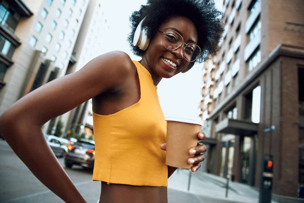 Pretty black woman wearing headphones while walking down the street listening to music drinking hot tea.