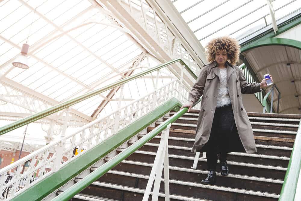 Black woman with curly tresses rushing downstairs with her phone in hand.