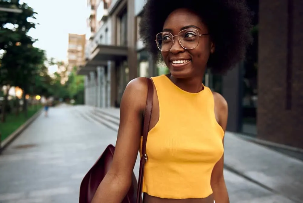 A joyful woman in a casual yellow shirt carrying a bag as she strolls through the city wearing glasses.