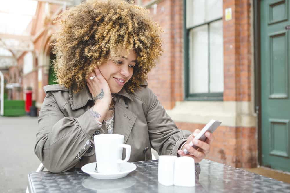 A woman with a beautiful curly hair type sitting outside at a cafe with a cup of coffee.