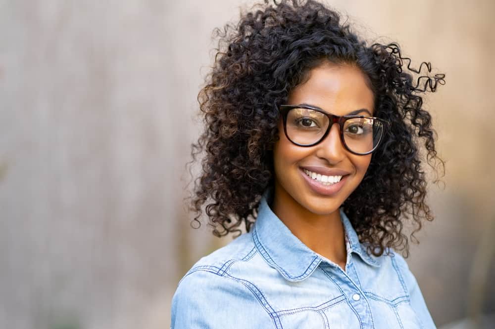 A cute young African American female is smiling while wearing black eyeglasses, a denim shirt, and neutral lipstick.