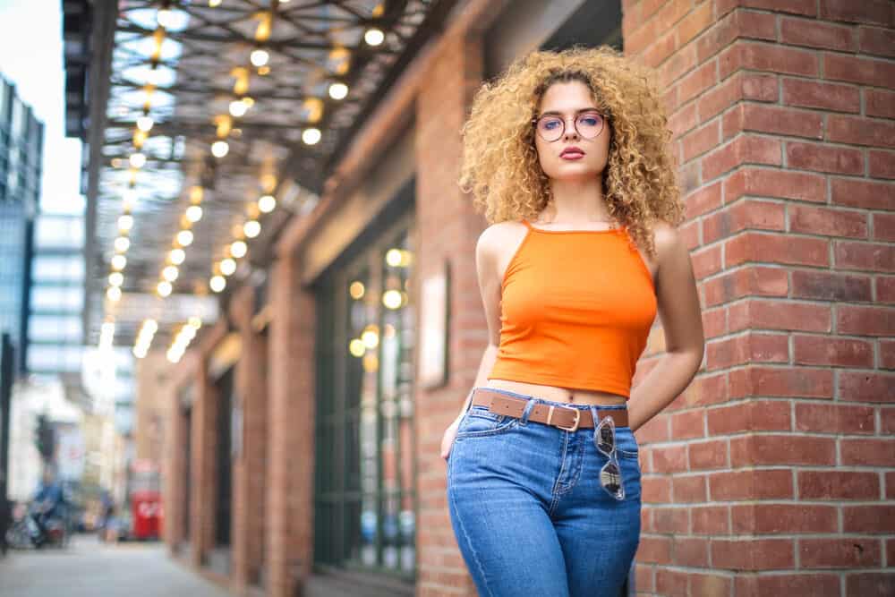 White female wearing an orange shirt and blue jeans with wavy hair with orange tones and underlying pigments