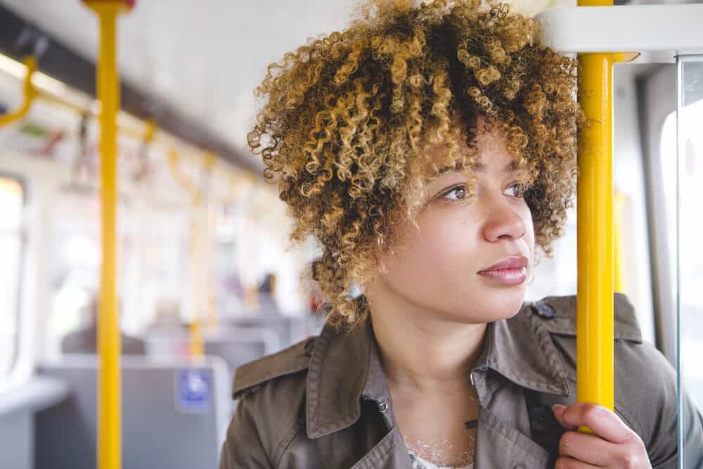 A woman with type 3 hair strands riding the subway is shown from a close-up while gripping the handrails and looking out of the window.