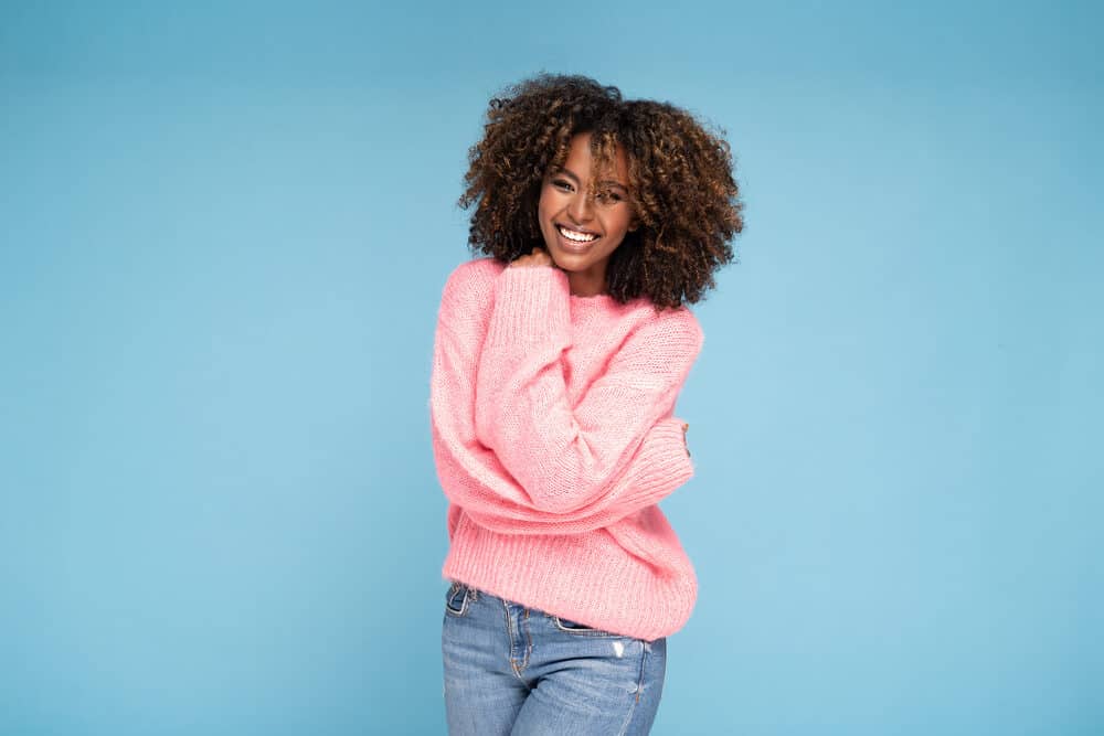 Black girl with curly wild tresses styled with coconut milk wearing a pink sweater and blue jeans.