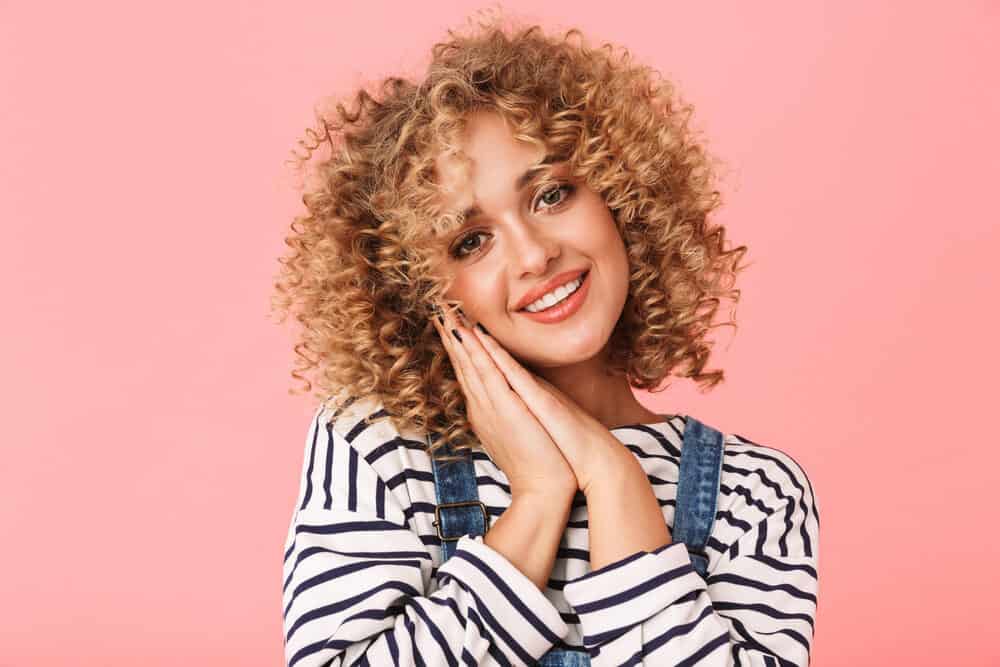 White woman with naturally curly hair with a deva haircut smiling wearing blue jean overalls