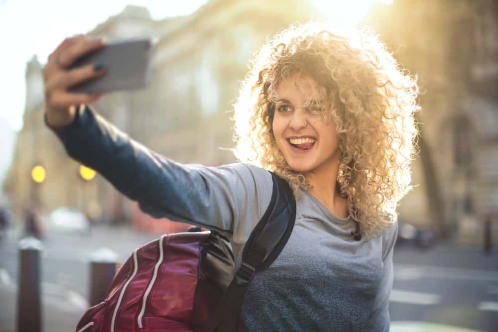 Ein schönes Mädchen macht ein tolles Selfie von sich mit dunkleren Wurzeln und blonden Wellen.