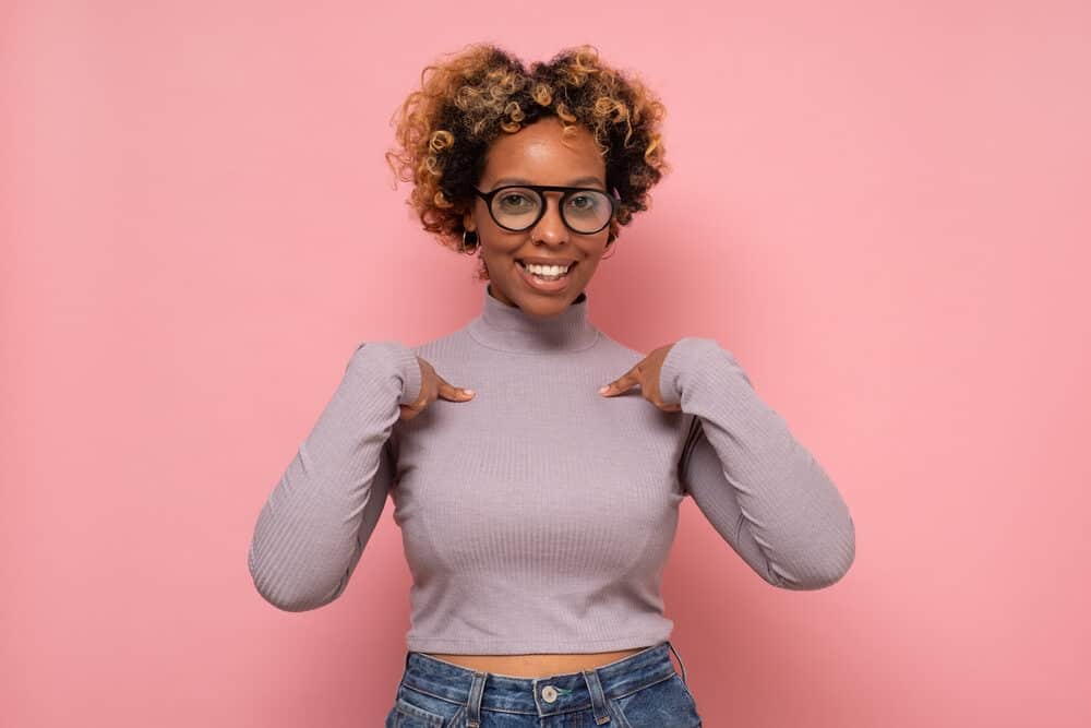 Black girl pointing fingers towards herself wearing a gray shirt with yellow brassy tones within her hair