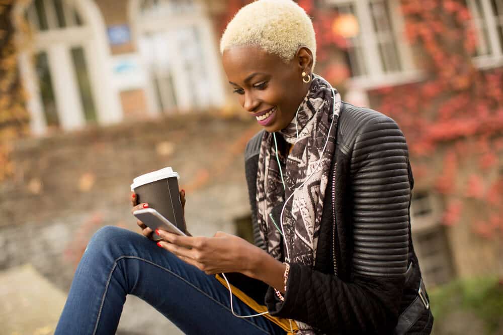 An attractive African American female with blue eyes sitting near the beach watching YouTube on her iPhone