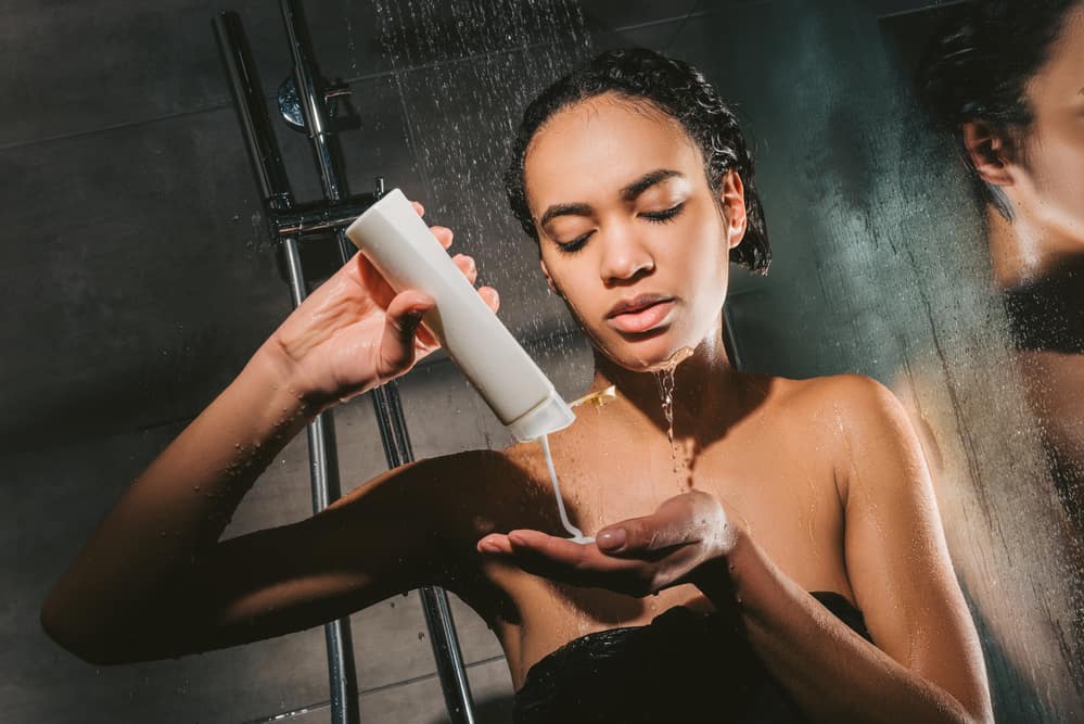 Black girl holding a body wash bottle with a milky texture shower gel while standing in the shower
