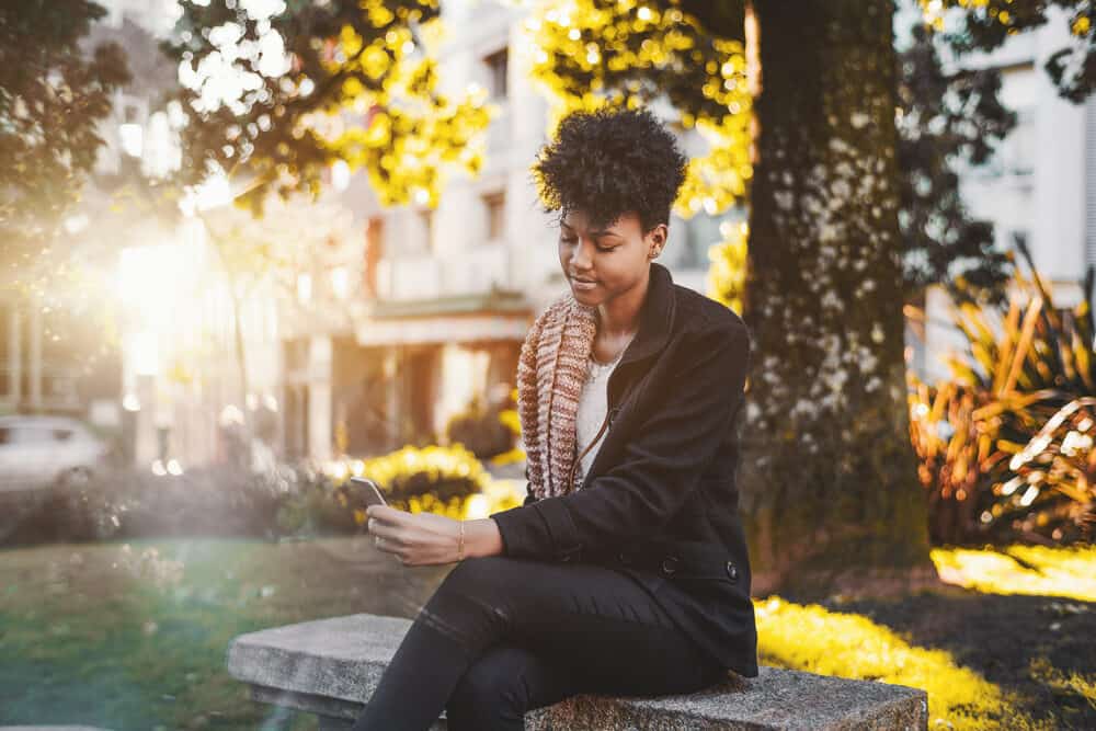 Young Brazilian female in warm clothes sitting on a park bench having a video call with her family