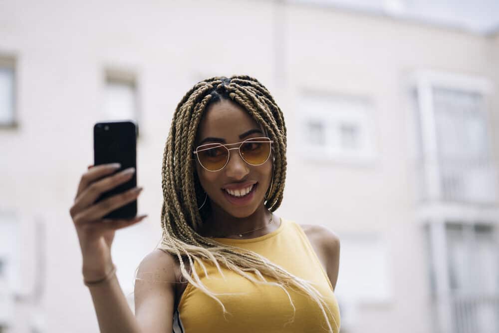 Latin female with fresh braids wearing sunglasses and a yellow dress while using a mobile phone