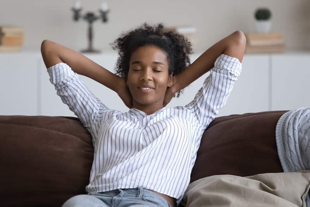 Peaceful African American woman with a curly hair type resting on her couch with her arms folded behind her head