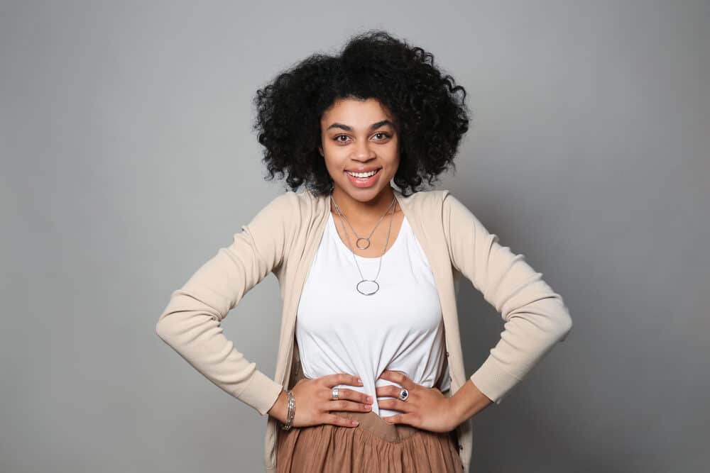 Black women with naturally curly hair wearing a light brown sweater, white shirt, brown skirt, and a ring necklace.