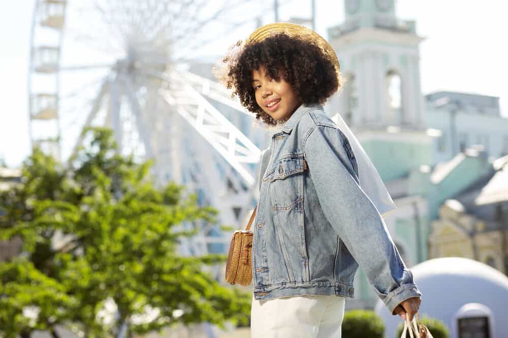 A woman with curly hair in a straw hat and denim jacket is shown carrying goods.