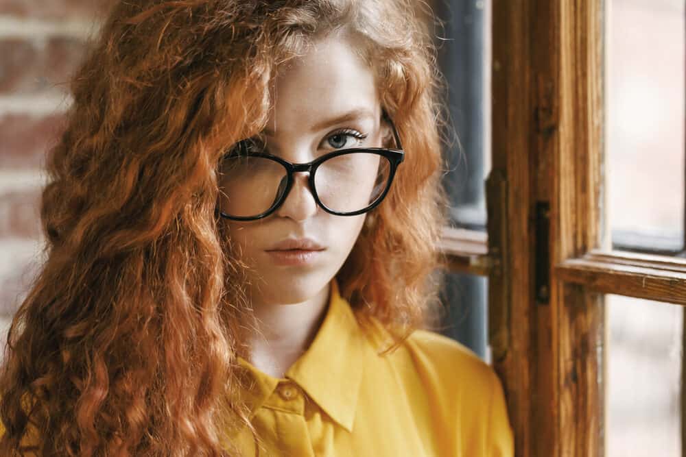Girl with ginger highlighted hair standing near a window wearing a yellow dress shirt and black eyeglasses.